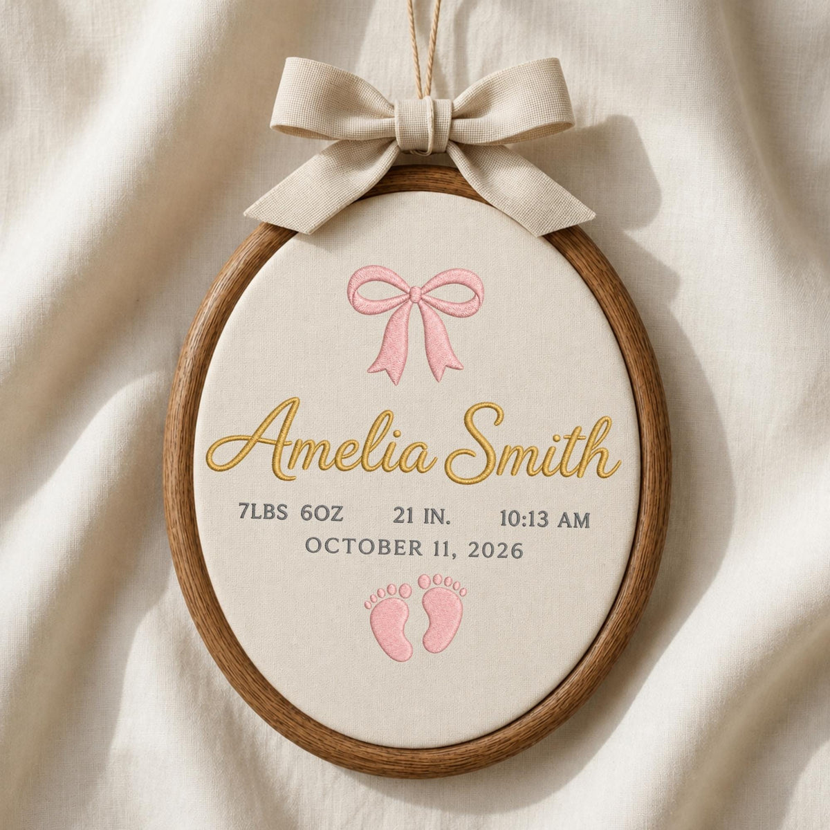 May include: An oval wooden embroidery hoop containing cream fabric with a beige ribbon bow at the top. Embroidered text reads Amelia Smith, 7lbs 6oz, 21 in., 10:13 AM, October 11, 2026, with a pink bow icon and two small pink baby footprints.