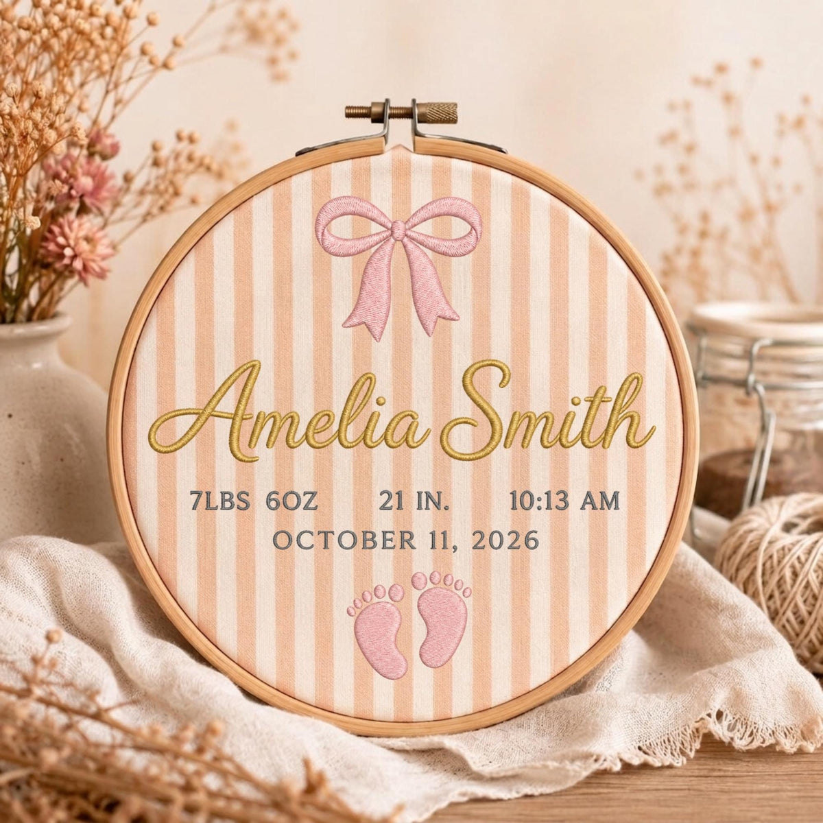 May include: A circular wooden embroidery hoop containing fabric with vertical peach and white stripes. The fabric features an embroidered pink bow, the name Amelia Smith in gold script, and birth details: 7lbs 6oz, 21 in, 10:13 am, October 11, 2026, and two small pink baby footprints.
