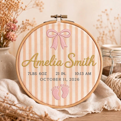 May include: A circular wooden embroidery hoop containing fabric with vertical peach and white stripes. The fabric features an embroidered pink bow, the name Amelia Smith in gold script, and birth details: 7lbs 6oz, 21 in, 10:13 am, October 11, 2026, and two small pink baby footprints.