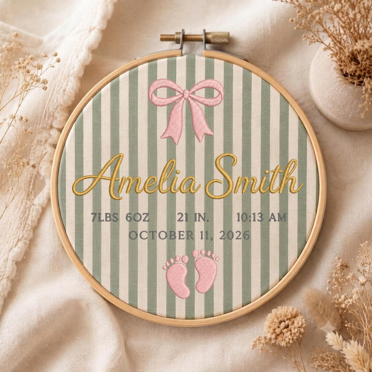 May include: A circular wooden embroidery hoop containing fabric with sage green and white vertical stripes. It features an embroidered pink bow, the name Amelia Smith in gold script, and birth details: 7lbs 6oz, 21 in, 10:13 am, October 11, 2026, and two pink baby footprints.