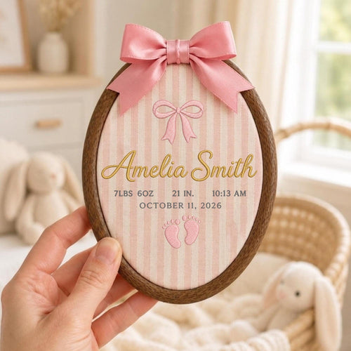 May include: An oval wooden embroidery hoop containing fabric with pink vertical stripes. It features a pink satin bow at the top, embroidered text reading Amelia Smith, 7lbs 6oz, 21 in, 10:13 am, October 11, 2026, and a pair of baby footprints.
