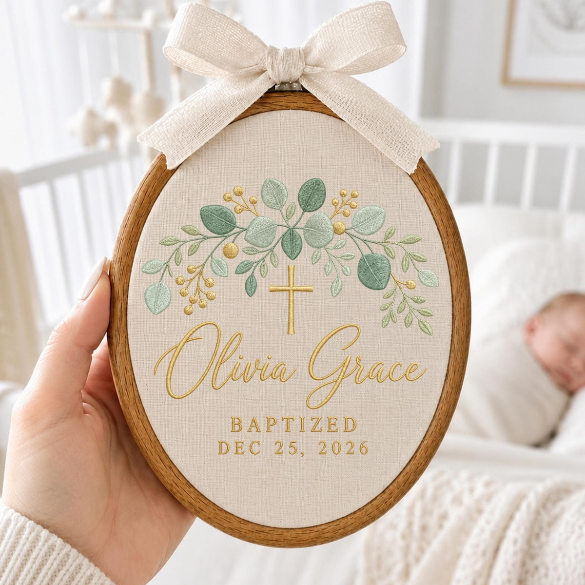 May include: An oval wooden embroidery hoop containing beige fabric with a green and gold embroidered eucalyptus wreath, a gold cross, and the text Olivia Grace, BAPTIZED, and DEC 25, 2026. A cream-colored fabric bow is attached to the top of the hoop.