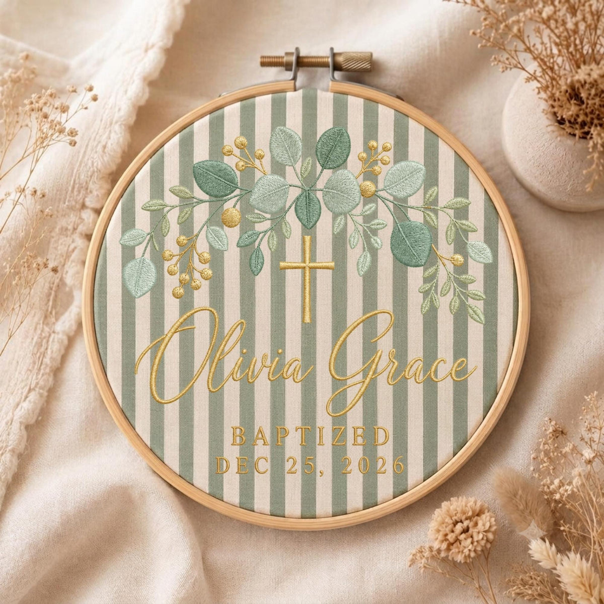 May include: A circular wooden embroidery hoop containing fabric with vertical sage green and white stripes. The embroidery features a gold cross, green eucalyptus leaves, gold berries, and the text Olivia Grace Baptized Dec 25, 2026.