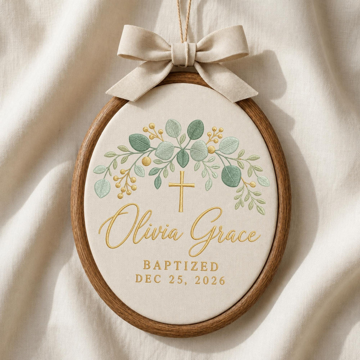 May include: An oval wooden embroidery hoop frame containing cream fabric with a beige ribbon bow at the top. The fabric features a green eucalyptus leaf design, a gold cross, and the text Olivia Grace, BAPTIZED, DEC 25, 2026 in gold script.