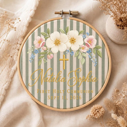 May include: A round wooden embroidery hoop containing fabric with sage green and white vertical stripes. The fabric features embroidered white and pink flowers, a gold cross, and the text Natalia Sophia First Holy Communion Sept 11, 2026.