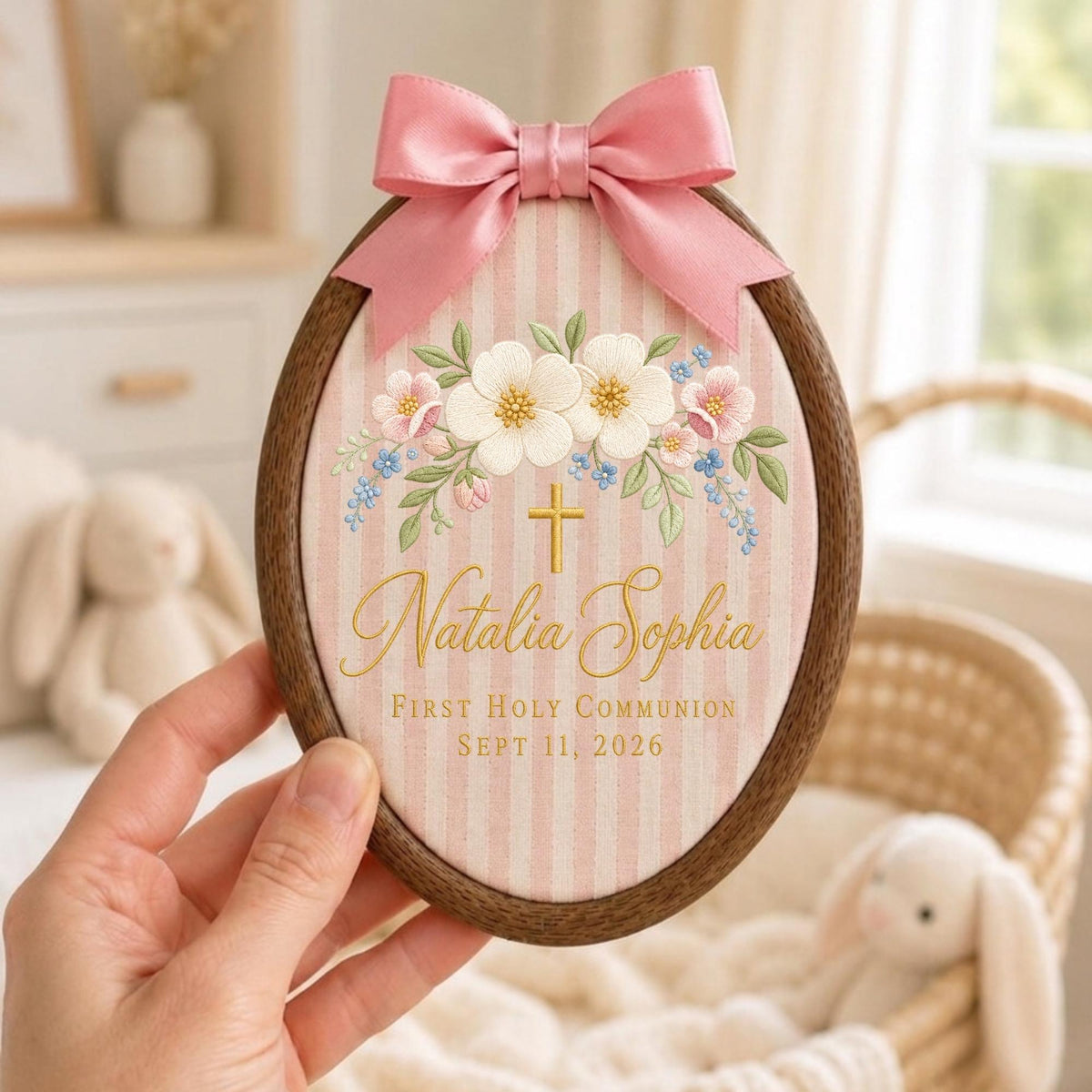 May include: An oval wooden embroidery hoop containing pink and white striped fabric with a floral arrangement, a gold cross, and the text Natalia Sophia First Holy Communion Sept 11, 2026. A pink satin ribbon bow is attached to the top of the frame.