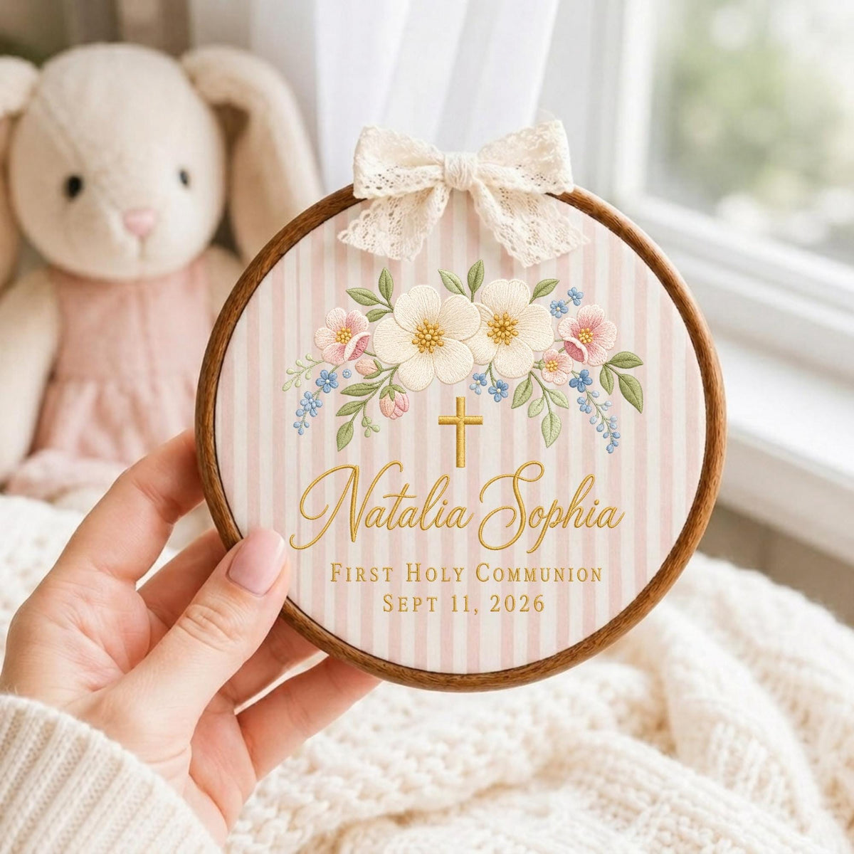 May include: A round wooden embroidery hoop frame containing fabric with pink and white vertical stripes. The fabric features a floral arrangement, a gold cross, and the text "Natalia Sophia First Holy Communion Sept 11, 2026" with a lace bow.