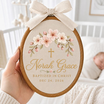 May include: An oval wooden embroidery hoop containing beige fabric with a floral design of pink cherry blossoms, a gold cross, and the embroidered text Norah Grace, BAPTIZED IN CHRIST, and DEC 24, 2026. A beige fabric bow is attached to the top of the hoop.