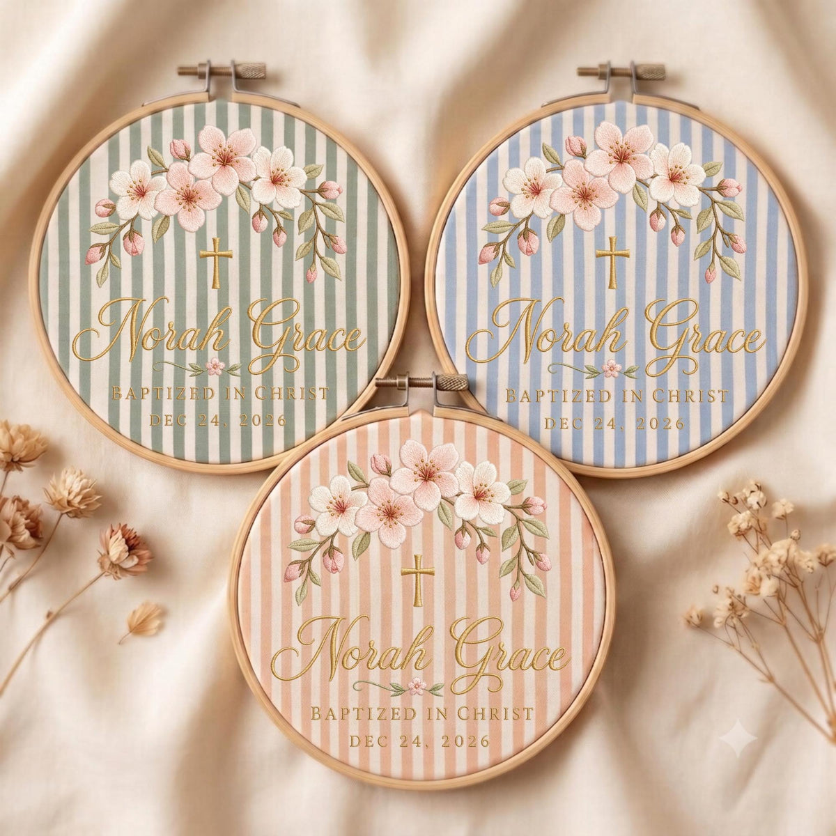 May include: Three circular wooden embroidery hoops featuring fabric with vertical stripes in sage green, light blue, and peach. Each displays a floral arch, a gold cross, and the text Norah Grace, Baptized in Christ, and Dec 24, 2026.