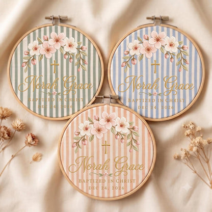 May include: Three circular wooden embroidery hoops featuring fabric with vertical stripes in sage green, light blue, and peach. Each displays a floral arch, a gold cross, and the text Norah Grace, Baptized in Christ, and Dec 24, 2026.