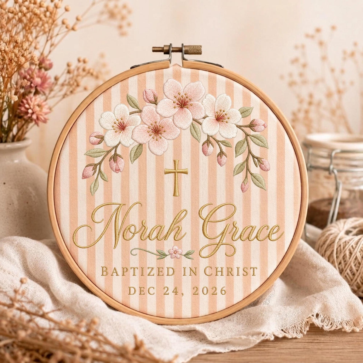 May include: A circular wooden embroidery hoop containing fabric with light peach vertical stripes. The embroidery features a floral arch of pink cherry blossoms, a gold cross, and the text Norah Grace, BAPTIZED IN CHRIST, DEC 24, 2026.