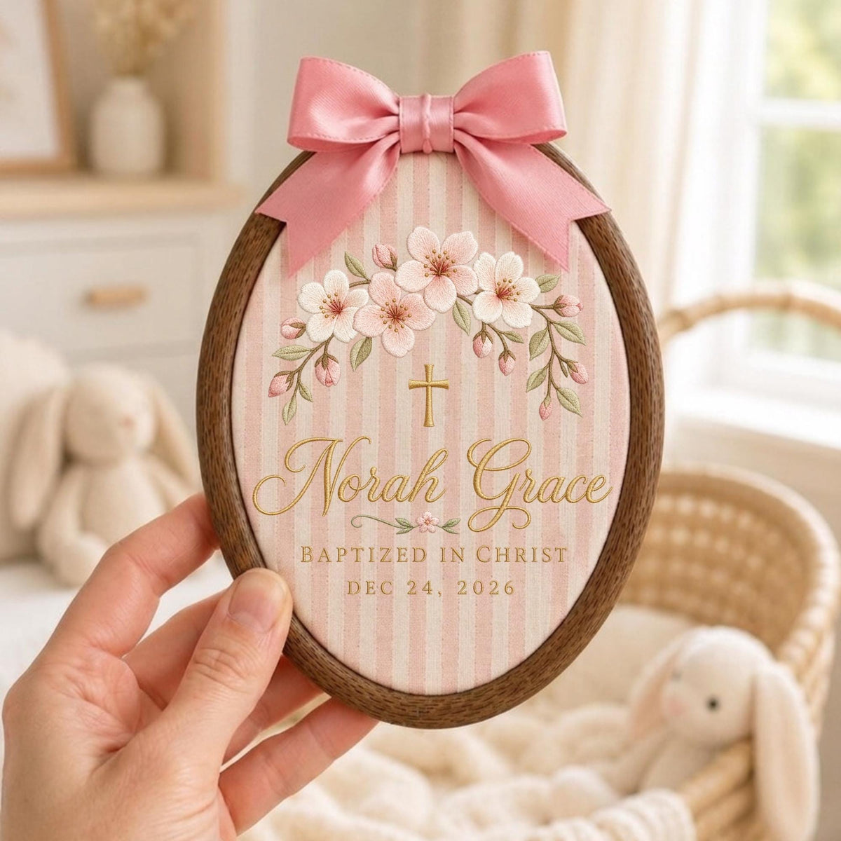 May include: An oval wooden embroidery hoop frame containing fabric with pink and white vertical stripes. The fabric features an embroidered floral arch, a gold cross, and the text Norah Grace BAPTIZED IN CHRIST DEC 24, 2026. A pink satin bow is attached to the top.