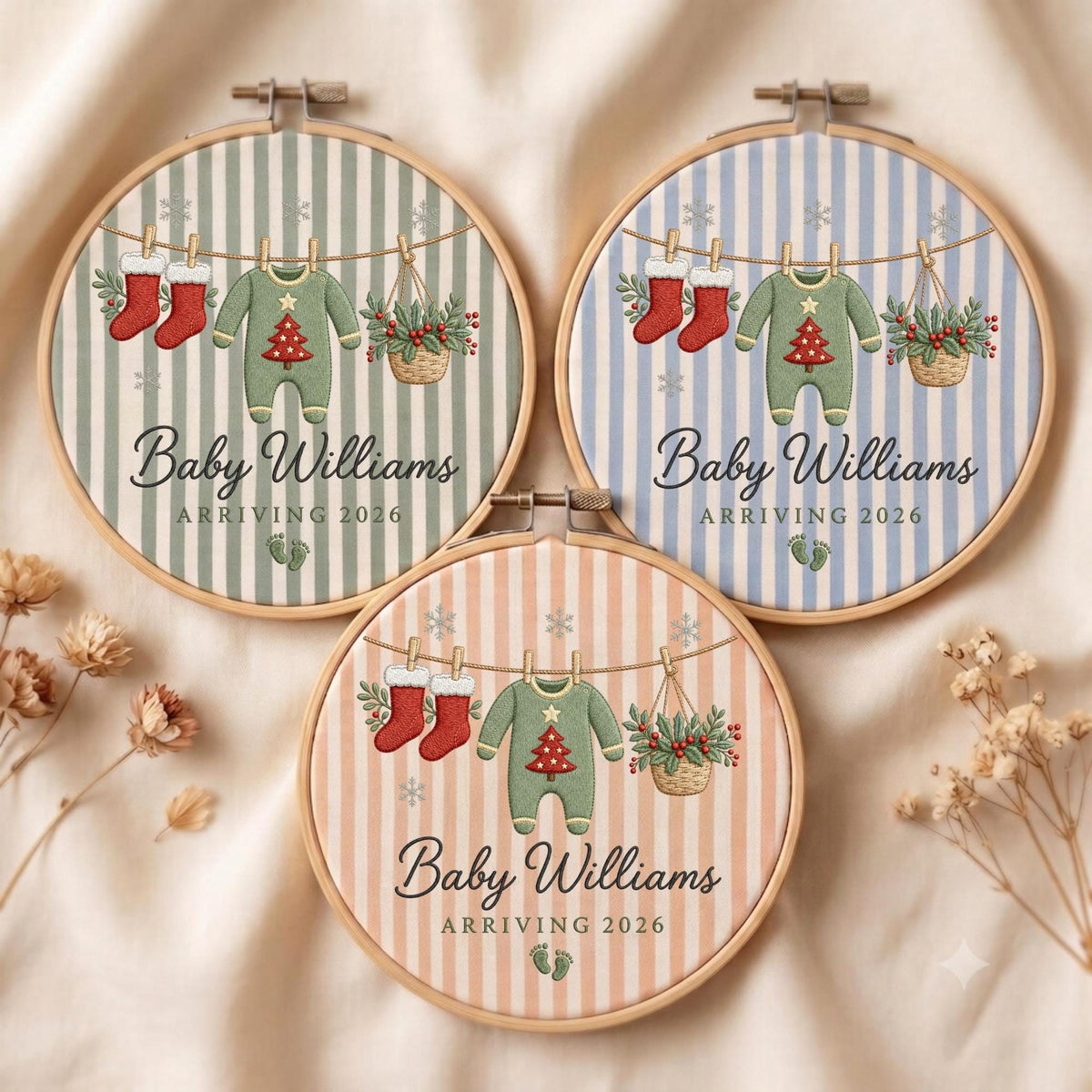 May include: Three circular wooden embroidery hoops featuring striped fabric backgrounds in sage green, light blue, and peach. Each displays a clothesline with two red stockings, a green baby onesie with a red Christmas tree, and a basket of holly. Text reads Baby Williams ARRIVING 2026 with small footprints.