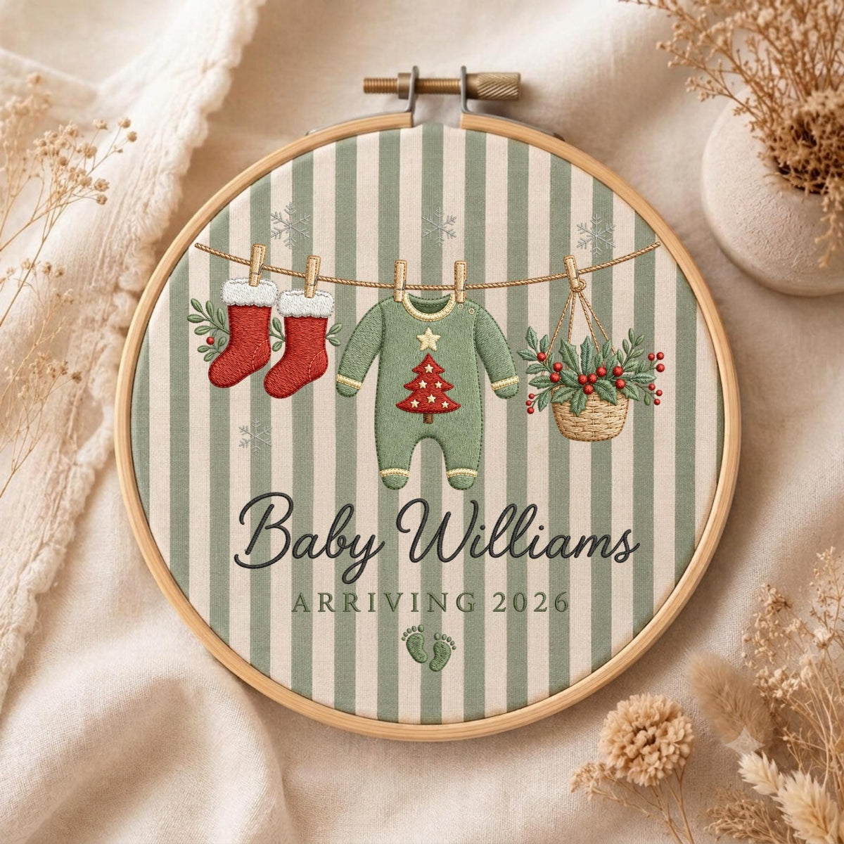 May include: A circular wooden embroidery hoop containing fabric with vertical sage green and white stripes. The embroidery features two red stockings, a green baby onesie with a red Christmas tree, and a basket of holly hanging on a line. Text reads Baby Williams ARRIVING 2026 with two small footprints.