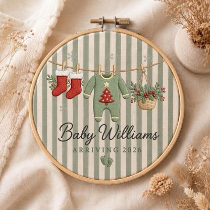 May include: A circular wooden embroidery hoop containing fabric with vertical sage green and white stripes. The embroidery features two red stockings, a green baby onesie with a red Christmas tree, and a basket of holly hanging on a line. Text reads Baby Williams ARRIVING 2026 with two small footprints.