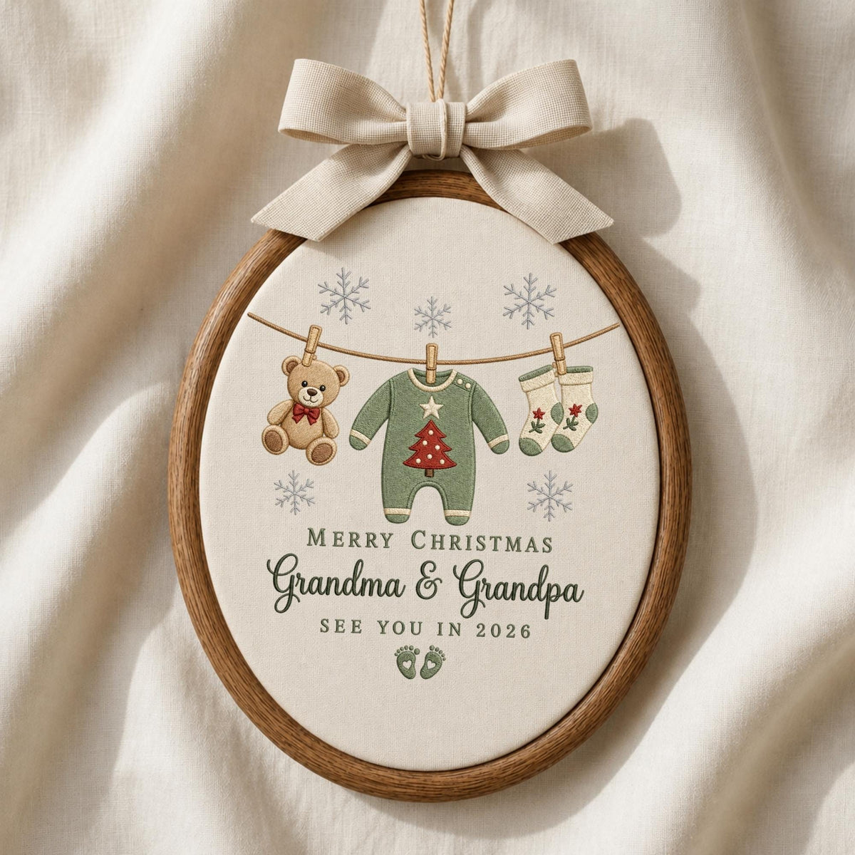 May include: An oval embroidery hoop ornament featuring a beige fabric background with a brown wooden frame and a beige ribbon bow. The design shows a teddy bear, a green onesie with a red tree, and socks hanging on a line with snowflakes. Text reads: MERRY CHRISTMAS Grandma &amp; Grandpa SEE YOU IN 2026 with two small footprints.