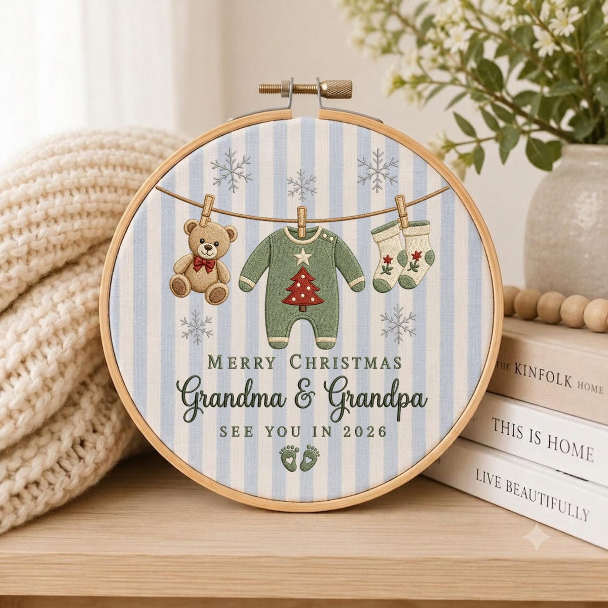 May include: A round wooden embroidery hoop containing fabric with light blue vertical stripes and embroidered snowflakes. It features a teddy bear, a green baby onesie with a red tree, and socks hanging on a line, with text reading Merry Christmas Grandma &amp; Grandpa See You In 2026 and baby footprints.