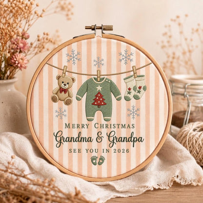 May include: A circular wooden embroidery hoop featuring a light pink and white striped fabric background. The design includes a clothesline with a teddy bear, a green baby onesie with a red Christmas tree, and two socks. Text reads: MERRY CHRISTMAS GRANDMA &amp; GRANDPA SEE YOU IN 2026.