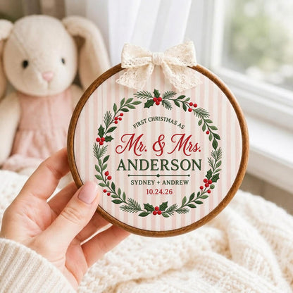 May include: A round wooden embroidery hoop containing fabric with a pink and white striped pattern. The fabric features a green holly wreath with red berries and the text: FIRST CHRISTMAS AS Mr. &amp; Mrs. ANDERSON SYDNEY + ANDREW 10.24.26. A cream lace bow is attached to the top.