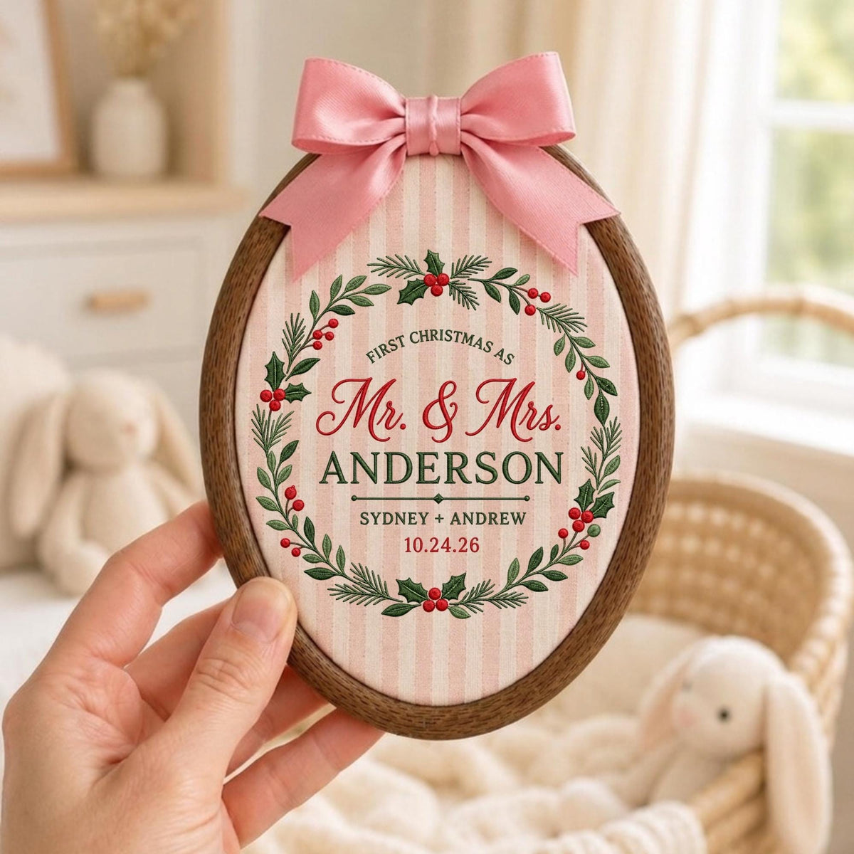 May include: An oval wooden embroidery hoop frame containing fabric with pink and white vertical stripes. A pink satin bow is attached to the top. The fabric features a green holly wreath with red berries and text reading First Christmas as Mr. &amp; Mrs. ANDERSON SYDNEY + ANDREW 10.24.26.
