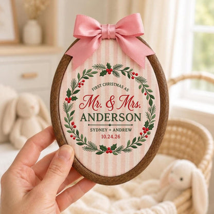 May include: An oval wooden embroidery hoop frame containing fabric with pink and white vertical stripes. A pink satin bow is attached to the top. The fabric features a green holly wreath with red berries and text reading First Christmas as Mr. &amp; Mrs. ANDERSON SYDNEY + ANDREW 10.24.26.