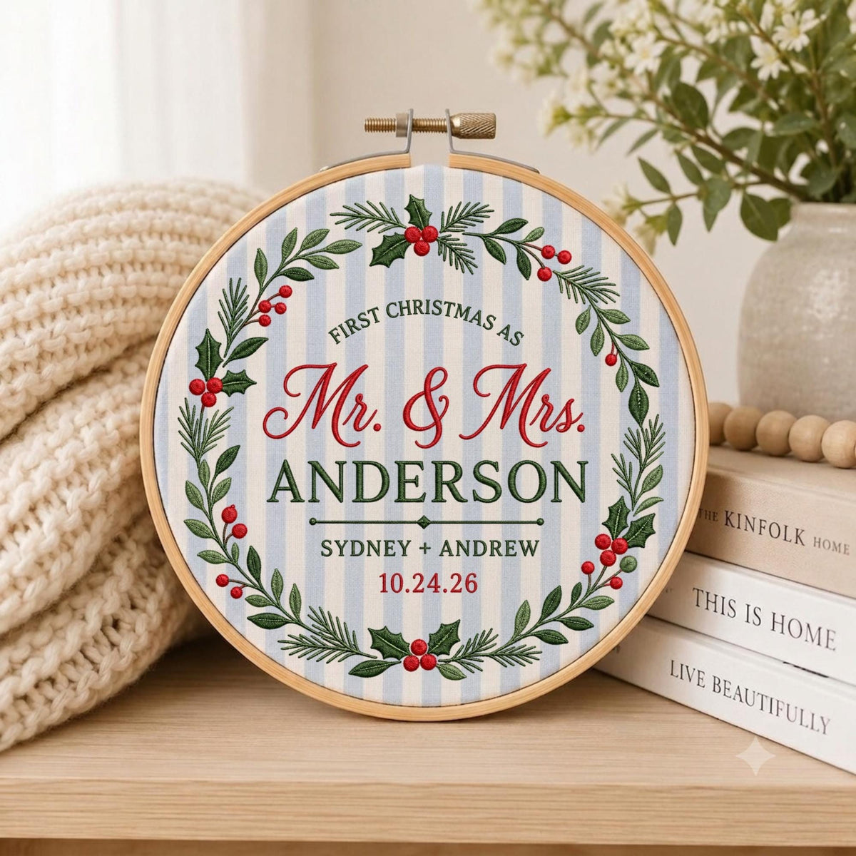 May include: A circular embroidery hoop containing fabric with light blue and white vertical stripes. The fabric features an embroidered wreath of green leaves and red berries surrounding the text: FIRST CHRISTMAS AS Mr. &amp; Mrs. ANDERSON SYDNEY + ANDREW 10.24.26.