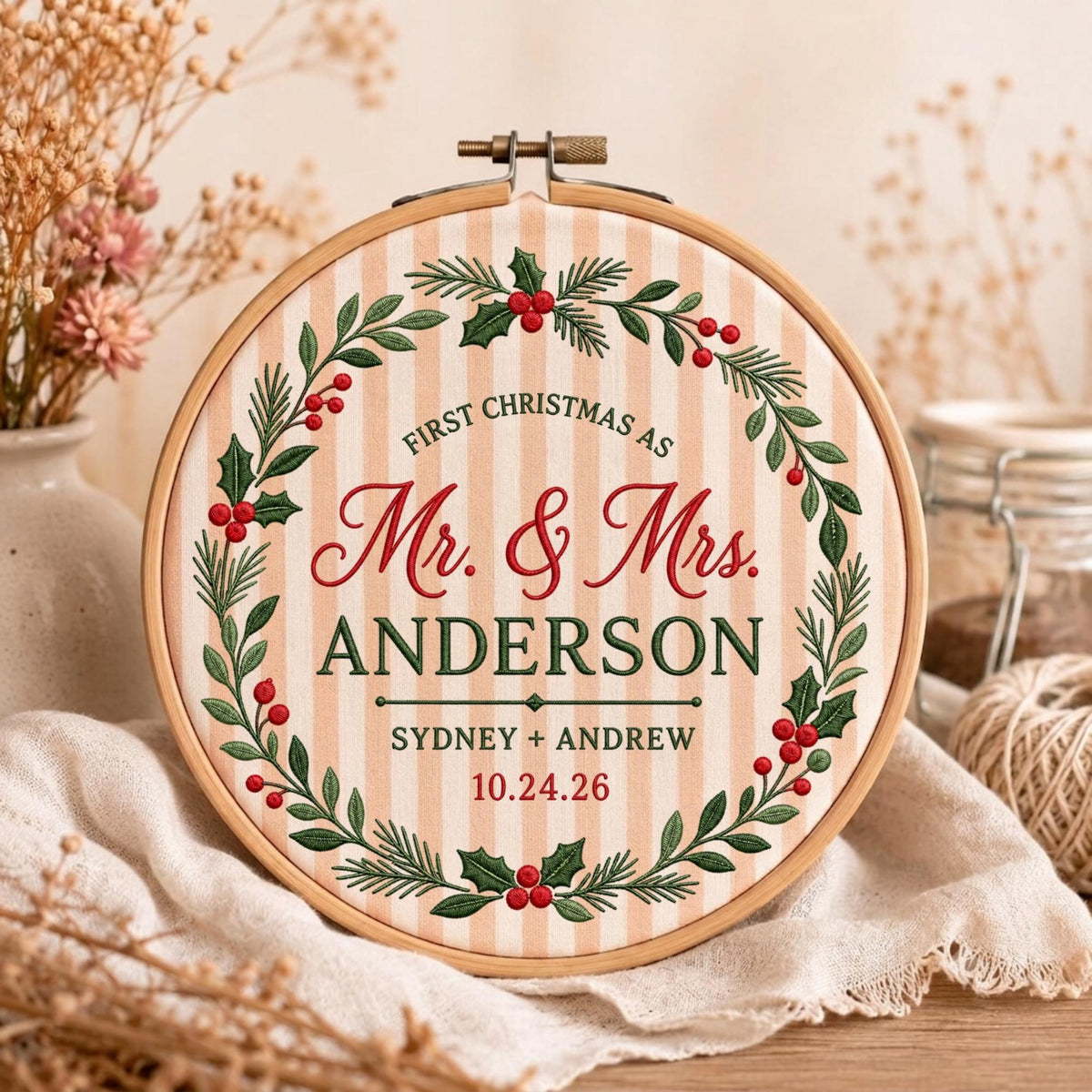 May include: A round wooden embroidery hoop containing fabric with vertical beige and white stripes. The fabric features an embroidered wreath of green holly leaves and red berries surrounding the text: FIRST CHRISTMAS AS Mr. &amp; Mrs. ANDERSON SYDNEY + ANDREW 10.24.26.