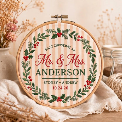 May include: A round wooden embroidery hoop containing fabric with vertical beige and white stripes. The fabric features an embroidered wreath of green holly leaves and red berries surrounding the text: FIRST CHRISTMAS AS Mr. &amp; Mrs. ANDERSON SYDNEY + ANDREW 10.24.26.