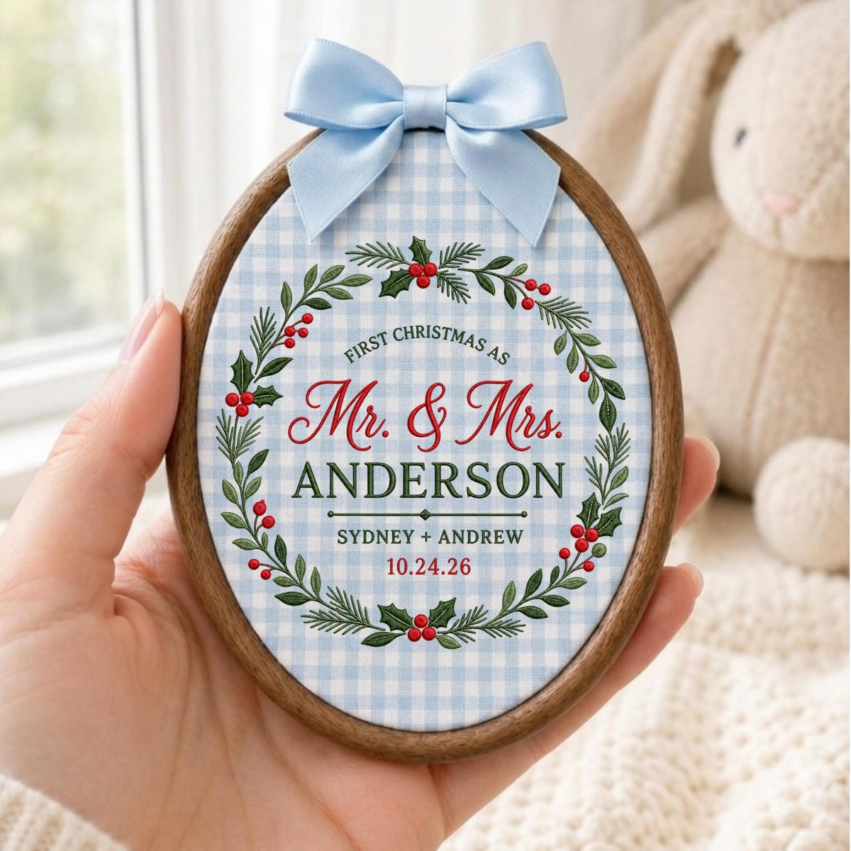 May include: An oval wooden embroidery hoop frame containing fabric with a light blue and white gingham pattern. A wreath of green pine needles and red berries surrounds the text: FIRST CHRISTMAS AS Mr. &amp; Mrs. ANDERSON SYDNEY + ANDREW 10.24.26.