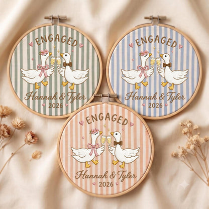 May include: Three circular wooden embroidery hoops featuring striped fabric in sage green, light blue, and peach. Each hoop displays two white geese clinking champagne glasses, with the text ENGAGED, Hannah &amp; Tyler, and the year 2026 printed.