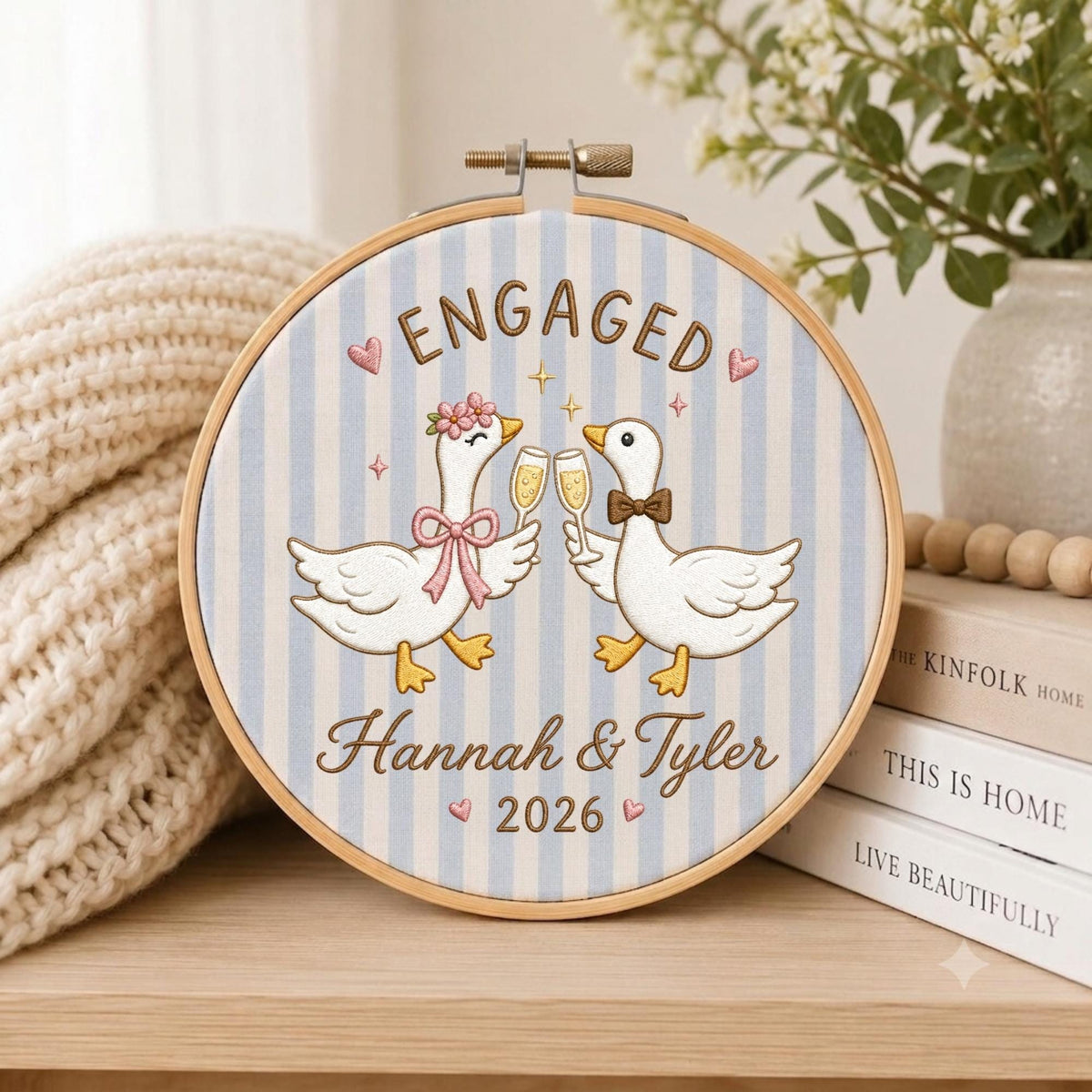 May include: A circular embroidery hoop featuring a light blue and white striped fabric background. Two white geese are illustrated in the center, holding champagne flutes. The text reads ENGAGED, Hannah &amp; Tyler, and 2026, accented by small pink hearts.
