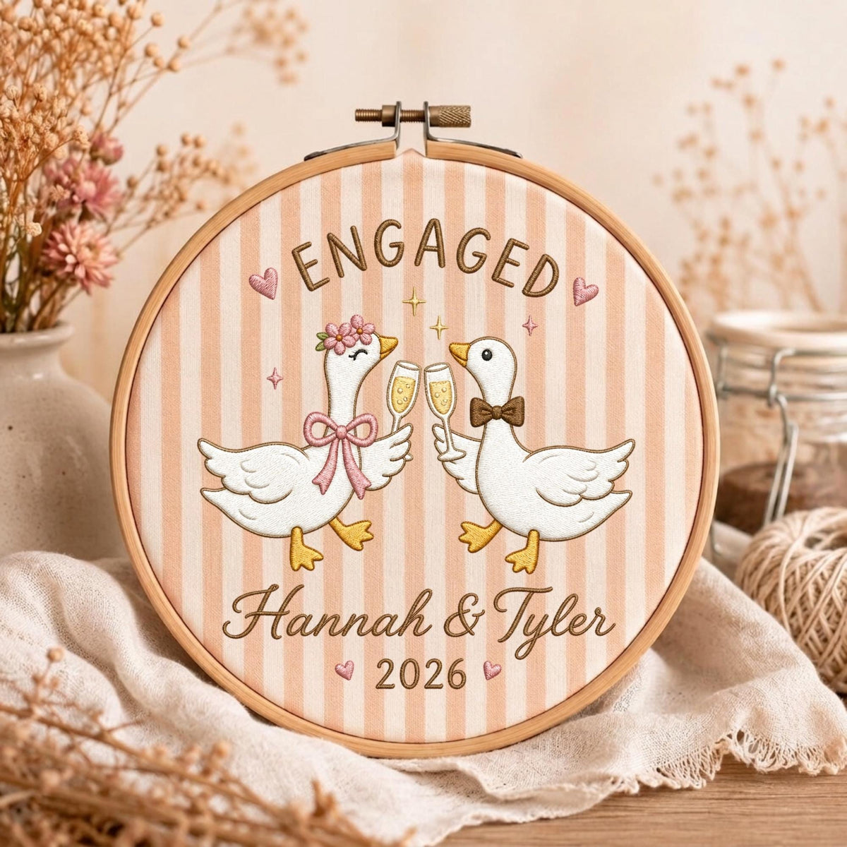 May include: A round wooden embroidery hoop containing fabric with vertical beige stripes. The embroidery features two white geese clinking champagne glasses, the text ENGAGED, the names Hannah &amp; Tyler, the year 2026, and small pink hearts.