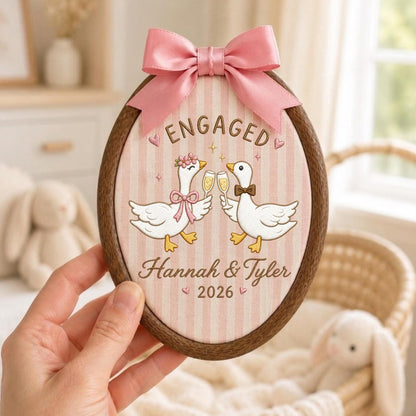 May include: An oval wooden embroidery hoop frame containing fabric with vertical pink stripes. The fabric features two white geese clinking champagne glasses, the text ENGAGED, Hannah &amp; Tyler, and 2026. A pink satin ribbon bow is attached at the top.