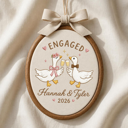 May include: An oval wooden embroidery hoop containing a cream-colored fabric with an embroidered design of two white geese clinking champagne glasses. The text reads ENGAGED, Hannah &amp; Tyler, and 2026. A beige fabric bow is attached to the top.