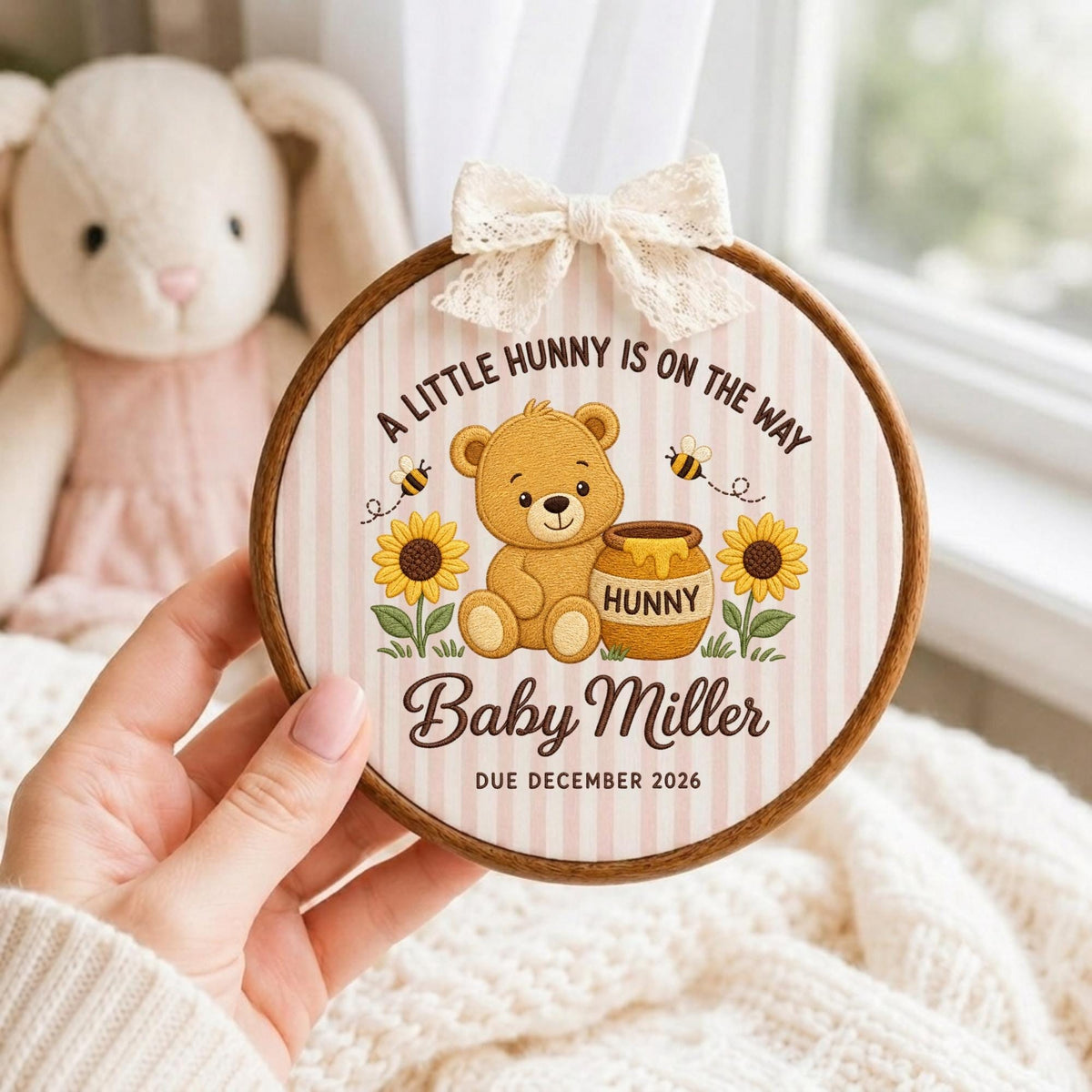 May include: A circular wooden embroidery hoop featuring a light pink and white striped fabric background. The design includes a brown teddy bear, a honey pot labeled HUNNY, sunflowers, bees, and the text A LITTLE HUNNY IS ON THE WAY, Baby Miller, and DUE DECEMBER 2026. A cream lace bow is attached to the top.