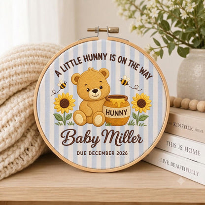 May include: A circular wooden embroidery hoop containing fabric with light blue vertical stripes. The design features a brown teddy bear, a honey pot labeled HUNNY, sunflowers, bees, and the text A LITTLE HUNNY IS ON THE WAY, Baby Miller, and DUE DECEMBER 2026.