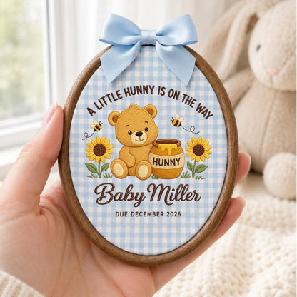May include: An oval wooden embroidery hoop featuring a light blue gingham fabric background. The design includes a brown teddy bear, a honey pot labeled HUNNY, sunflowers, and bees. Text reads: A LITTLE HUNNY IS ON THE WAY, Baby Miller, DUE DECEMBER 2026. A light blue satin bow is attached to the top.