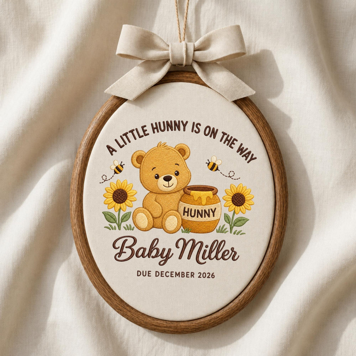 May include: An oval wooden embroidery hoop containing fabric with a printed design of a teddy bear, a honey pot labeled HUNNY, sunflowers, and bees. The text reads A LITTLE HUNNY IS ON THE WAY, Baby Miller, and DUE DECEMBER 2026. A beige bow is attached.