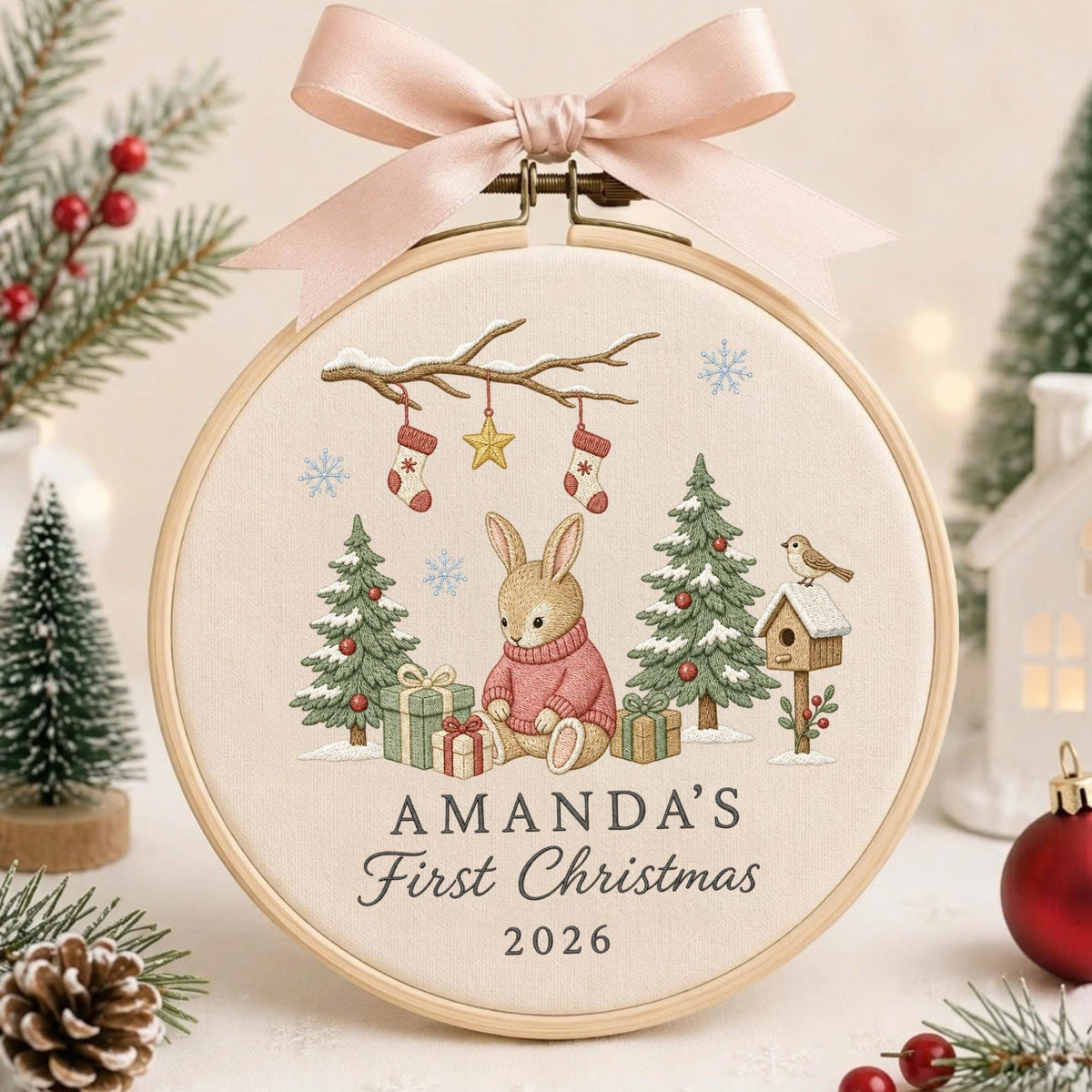May include: A round wooden embroidery hoop featuring a light-colored fabric with a Christmas-themed design. The embroidery depicts a rabbit in a pink sweater, pine trees, gifts, and a birdhouse. Text reads AMANDA'S First Christmas 2026. A pink ribbon is tied at the top.