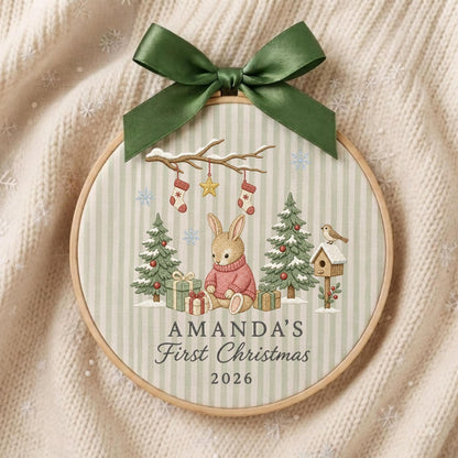 May include: A round wooden embroidery hoop featuring a light green and white striped fabric with a printed illustration of a rabbit in a pink sweater, Christmas trees, gifts, and stockings. The text reads AMANDA'S First Christmas 2026.