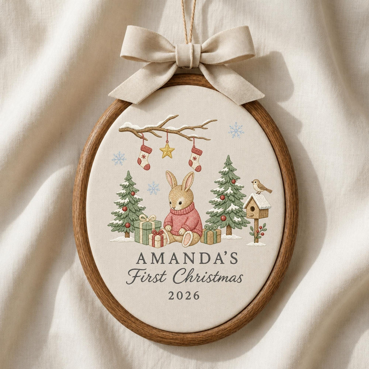 May include: An oval wooden embroidery hoop ornament featuring a beige fabric center with a printed illustration of a rabbit in a pink sweater, Christmas trees, gifts, and a birdhouse. The text reads AMANDA'S First Christmas 2026. A beige bow is attached.