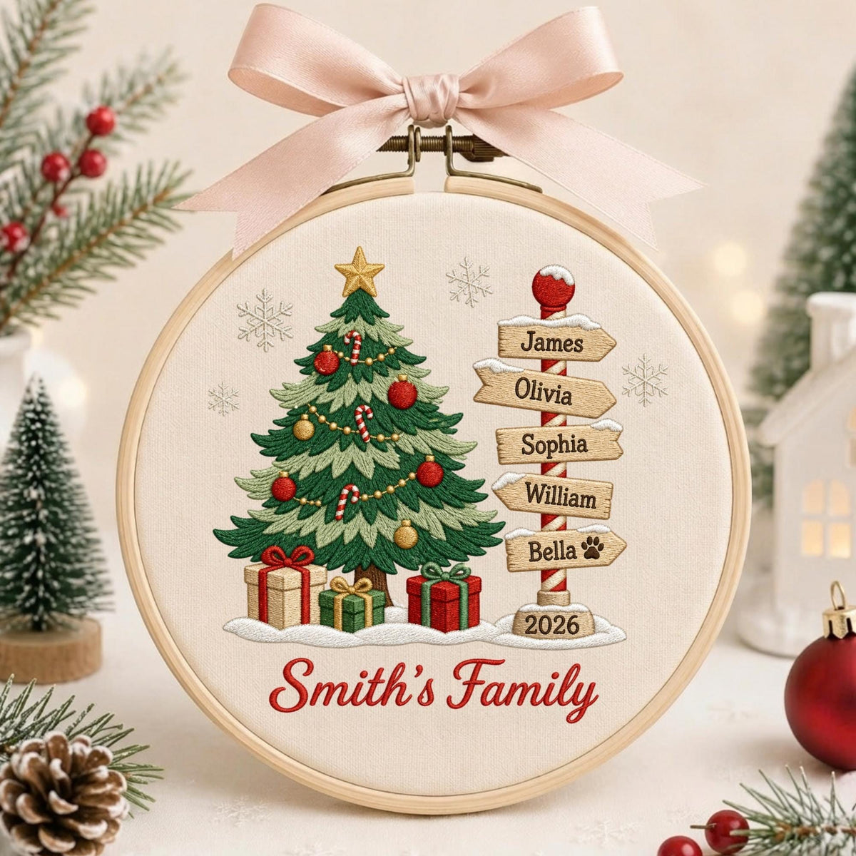 May include: A circular embroidery hoop featuring a stitched Christmas tree, wrapped gifts, and a signpost with the names James, Olivia, Sophia, William, and Bella, plus the year 2026. The text Smith's Family is embroidered below, topped with a pink bow.