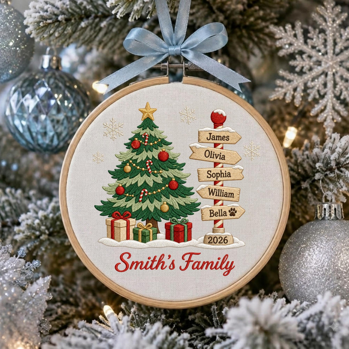 May include: A round wooden embroidery hoop ornament featuring a stitched Christmas tree with red ornaments and gifts, a signpost with the names James, Olivia, Sophia, William, and Bella, the year 2026, and the text Smith's Family, topped with a light blue ribbon.