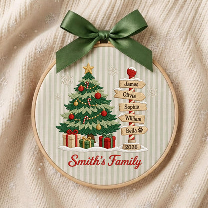 May include: A round embroidery hoop ornament featuring a green Christmas tree with ornaments, gifts, and a signpost with the names James, Olivia, Sophia, William, and Bella. It includes the text 2026 and Smith's Family, topped with a green satin bow.