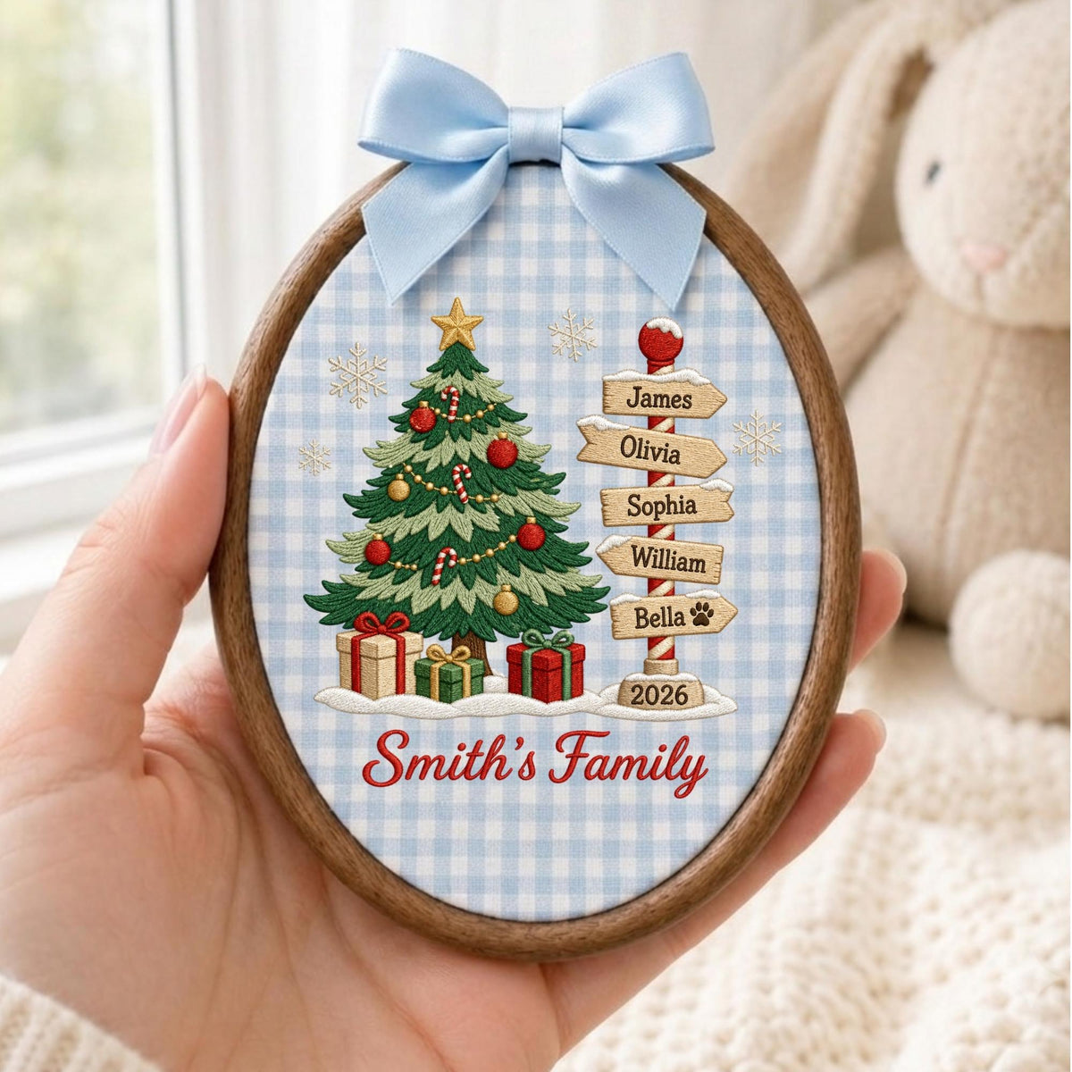 May include: An oval wooden embroidery hoop ornament featuring a light blue gingham fabric background. The design includes an embroidered Christmas tree with ornaments, gifts, a signpost with names James, Olivia, Sophia, William, Bella, and the year 2026.