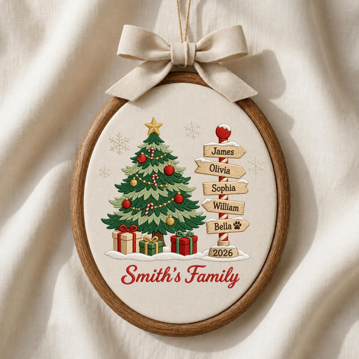 May include: An oval wooden embroidery hoop ornament featuring a cream fabric background. It displays a green Christmas tree with red and gold ornaments, gifts, and a signpost with the names James, Olivia, Sophia, William, and Bella. Text reads Smith's Family and 2026, topped with a beige fabric bow.