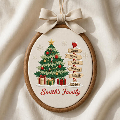May include: An oval wooden embroidery hoop ornament featuring a cream fabric background. It displays a green Christmas tree with red and gold ornaments, gifts, and a signpost with the names James, Olivia, Sophia, William, and Bella. Text reads Smith's Family and 2026, topped with a beige fabric bow.
