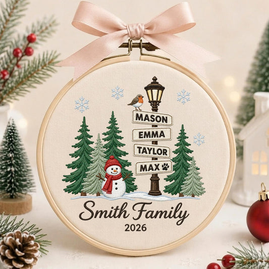 May include: A circular embroidery hoop featuring a cream fabric background with a winter scene. The design includes green pine trees, a snowman with a red scarf, a street sign with the names Mason, Emma, Taylor, Max, and the text Smith Family 2026.