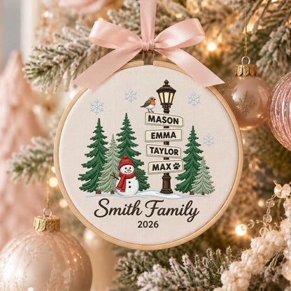 May include: A round wooden embroidery hoop ornament featuring a cream fabric background with a winter scene. It displays a snowman, green pine trees, a street lamp with signs reading Mason, Emma, Taylor, Max, and the text Smith Family 2026.