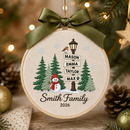 May include: A round embroidery hoop ornament featuring a cream fabric background with a winter scene. It displays green pine trees, a snowman with a red scarf, a street lamp with signs reading MASON, EMMA, TAYLOR, MAX, and Smith Family 2026.